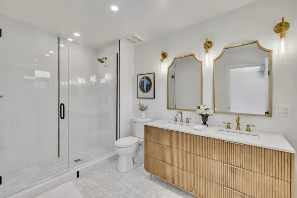 This modern bathroom in Phoenix, AZ features a double vanity with gold fixtures, a glass shower, and tasteful decor, captured with professional real estate photography.