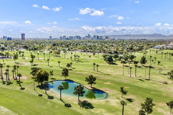 This aerial real estate photography captures a lush green golf course with a serene lake and palm trees in Phoenix, AZ, set against the stunning city skyline and mountain backdrop.