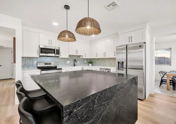 This modern kitchen in Mesa, AZ features a spacious island with black granite countertop, white cabinetry, stainless steel appliances, and stylish pendant lighting, captured by professional real estate photography.